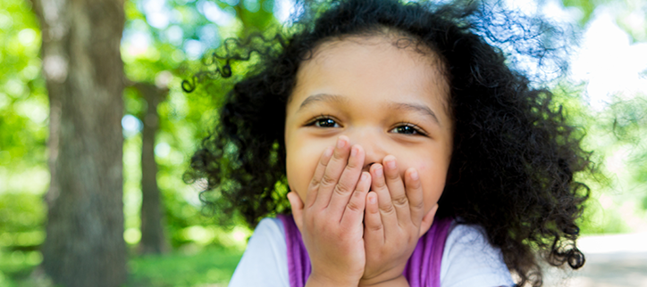 Little girl laughing and covering her mouth, standing in a forest.